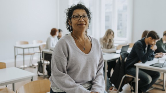 Kvindelig skolelærer sidder på bord og smiler med klasse i baggrunden